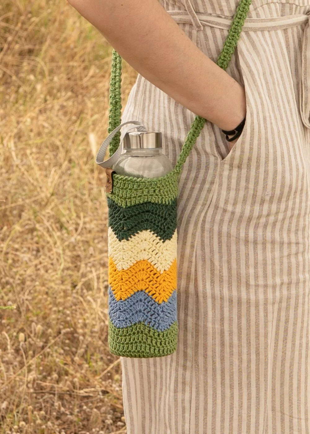 Funda de crochet hecha a mano para botella de agua con patrón de ondas en colores verde oliva, verde oscuro, amarillo mostaza, amarillo claro y azul claro, colgada en bandolera.