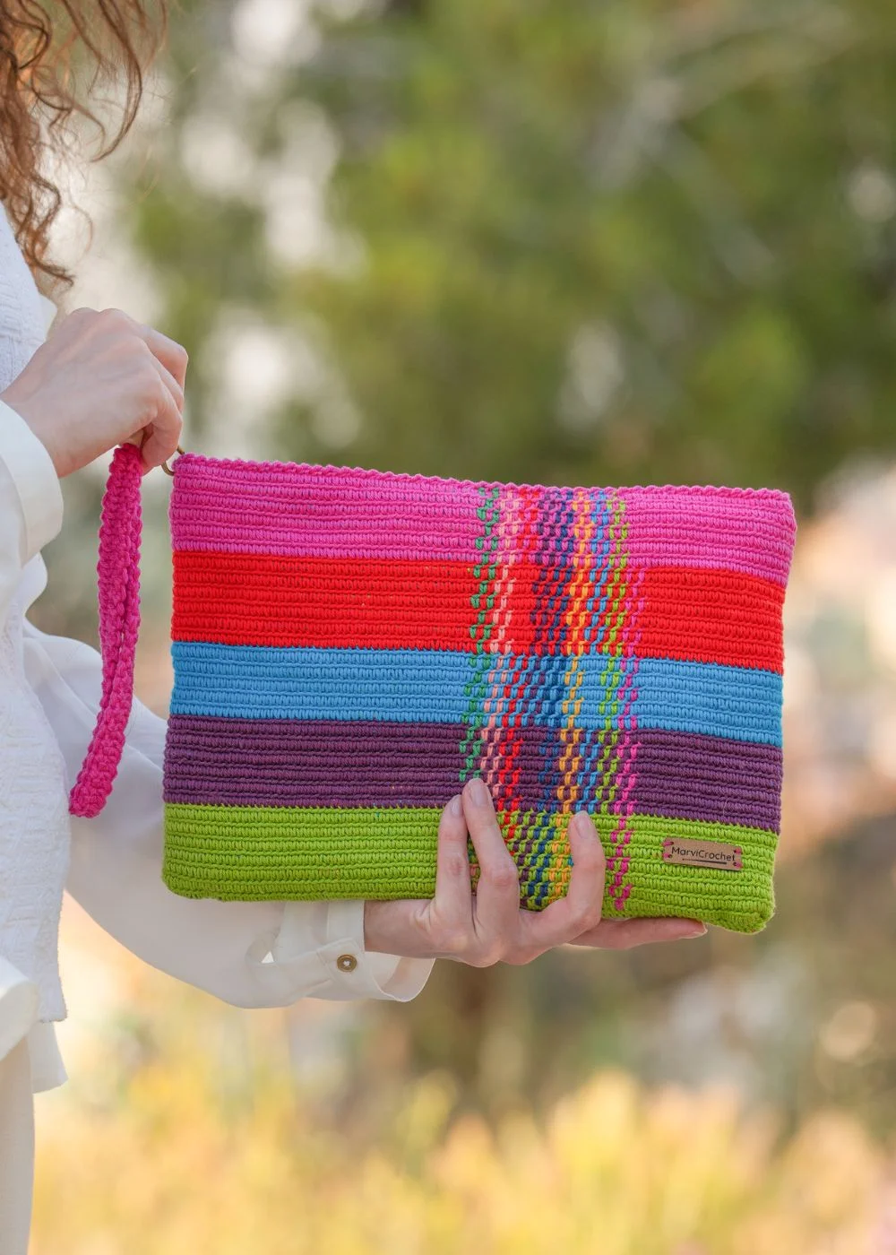 Bolso de mano de crochet con patrón de rayas vibrantes en tonos fucsia, rojo, azul turquesa, morado y verde, ideal para cualquier ocasión. Etiqueta Marvicrochet visible.