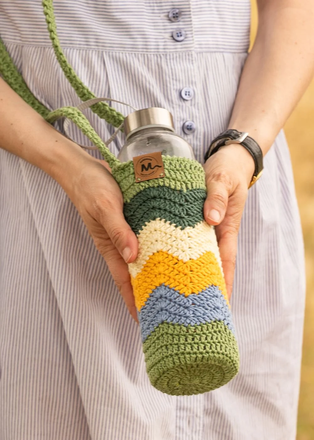 Funda de crochet hecha a mano para botella de agua con patrón de ondas en colores verde claro, verde oscuro, amarillo claro, amarillo mostaza y azul claro, colgada al hombro y sujetada con la mano.