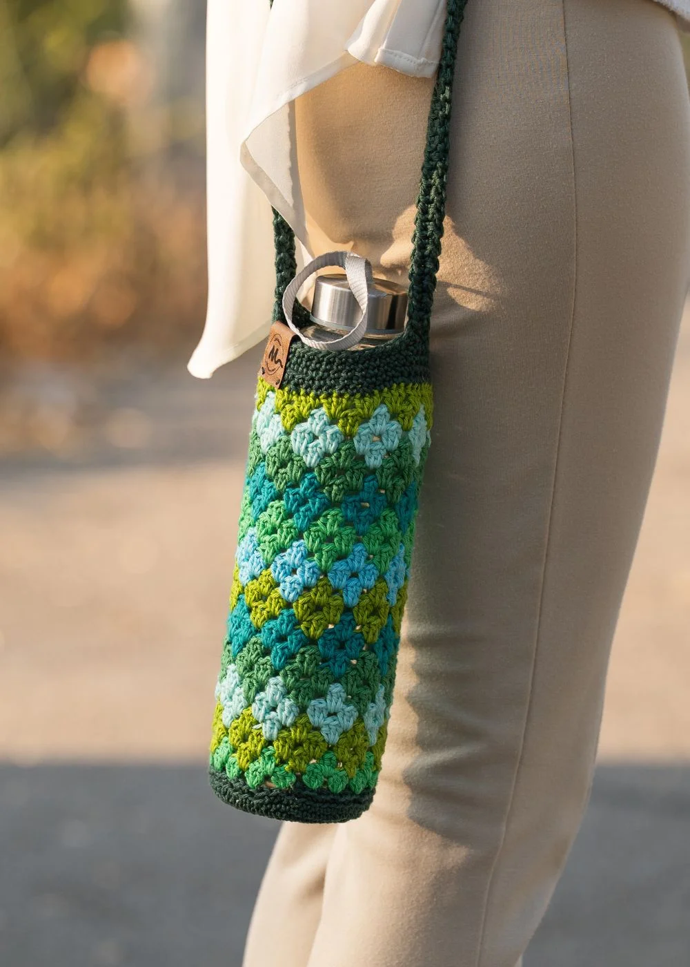 Funda de crochet artesanal para botella de agua en tonos verdes y azules, con patrón de rombos, colgada en el hombro.